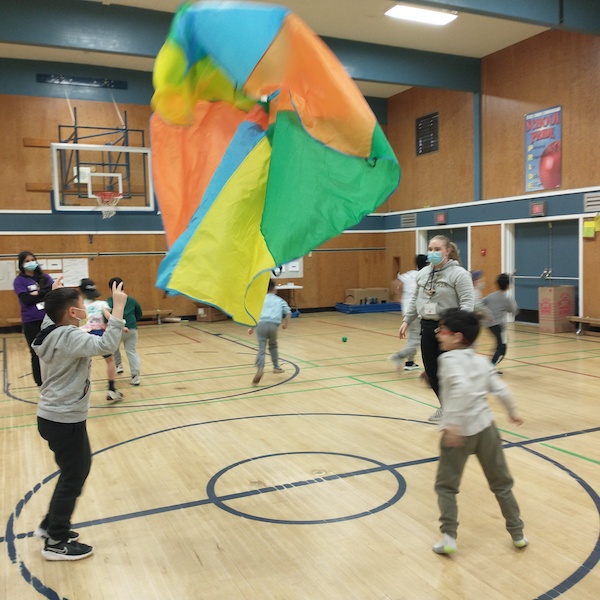 Kids playing parachute games at Spring Break, led by Taylor. They are in a circle and are watching the parachute, which has been thrown up into the air