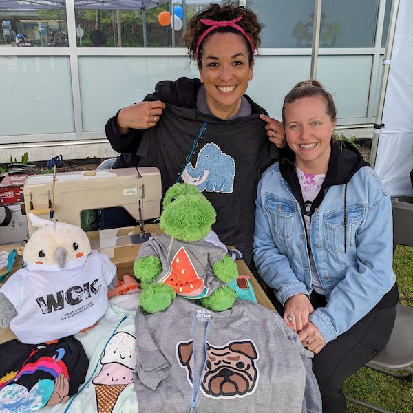 Mina and Kelsey at a WCK stall at an event, surrounded by Port Shirts and Mina's sewing machine. Mina is holding up a dark grey Port Shirt with a picture of a blue elephant on it.