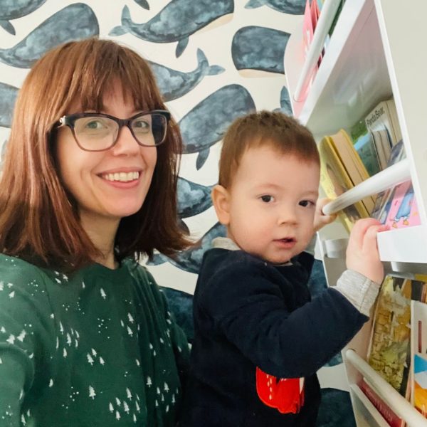 Mom Ashley and her baby Rhys smiling at the camera, each wearing PJs gifted by WCK. Rhys is picking a book from his bookshelf.