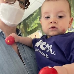 A baby in his mom's arms, holding rattle toys, wearing a purple WCK Port Shirt, looking at the camera. Mom is wearing a mask and glasses.