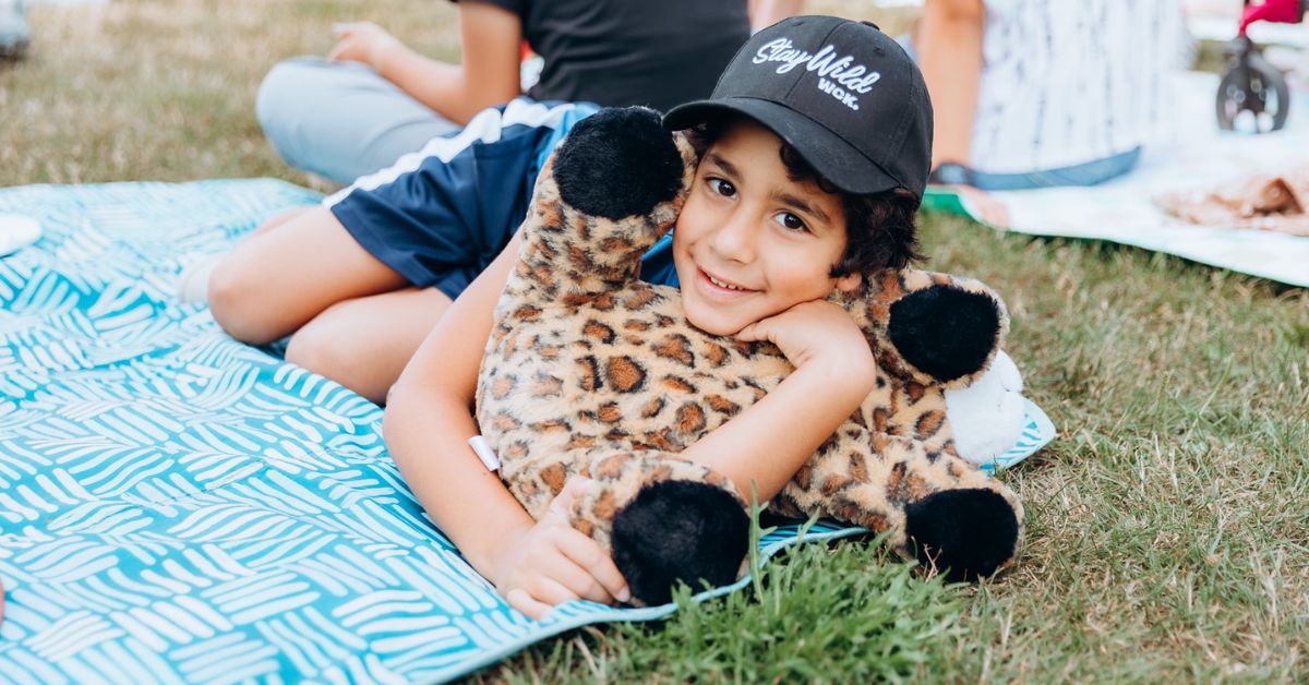 A child at City Camp wearing a WCK "Stay Wild" cap, lying on a blanket cuddling a stuffed toy giraffe and smiling.