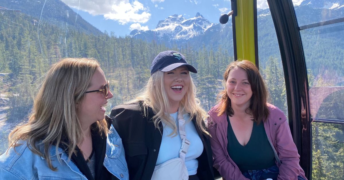 Jill (centre) with Kelsey and Hannah from the WCK team, sitting in the Sea to Sky Gondola in Squamish. There are mountains and trees in the background and it's a sunny day.
