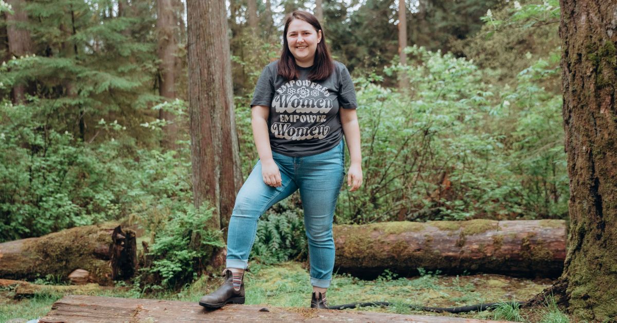 A photo of Abbey in a forest (taken at the 2023 She Can retreat). She has her right foot on a log and her hands by her side. She's smiling at the camera. Her t shirt says "Empowered women empower women"