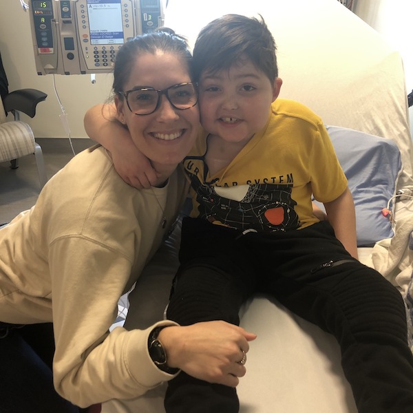 Cameron and nurse Carly, who both came up with the first ever WCK Port Shirts. Cameron is sitting on a hospital bed with his arm around Carly's shoulder, and they are smiling at the camera. Cameron is wearing a Port Shirt.