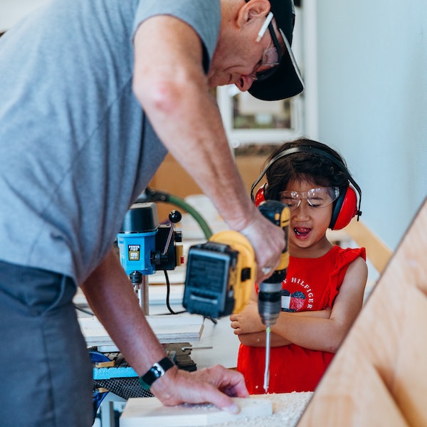 A girl at City Camp wearing goggles and ear defenders using power tools with Chuck's help