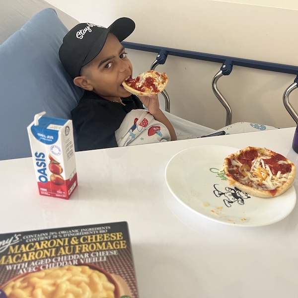 A 10-year-old boy in his hospital bed, eating a pizza from the WCK freezer and looking happily at the camera. On the bedside table is another small pizza that has been cooked, a box of mac and cheese (unopened), and a juice box. The boy is wearing a WCK baseball cap that says "Stay wild."