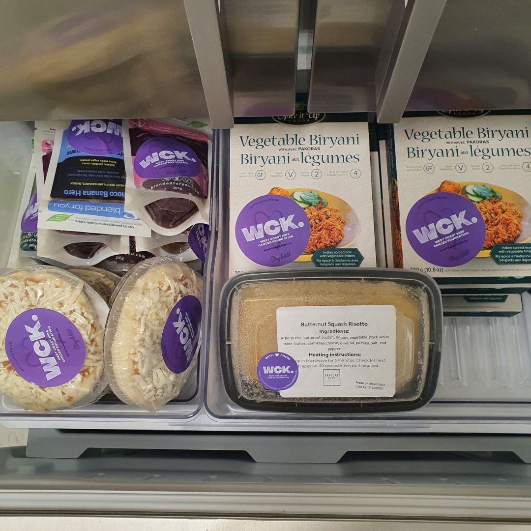 A photo of meals in one of the freezer drawers. Each meal has a purple WCK sticker on it. The meals include vegetable biryani, pizza, nutritious smoothies, and mashed potato.