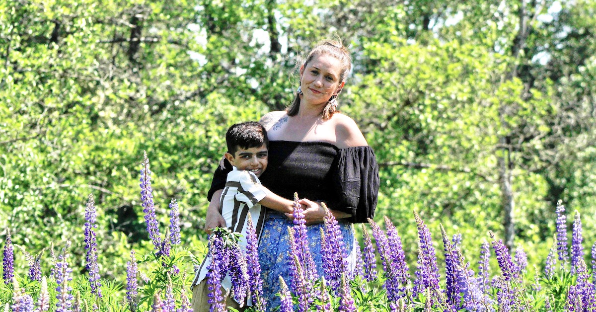 Ezra and his mum Amanda hugging, smiling at the camera. They are standing in front of green trees and there are lavender plants in front of them. It's a sunny day.