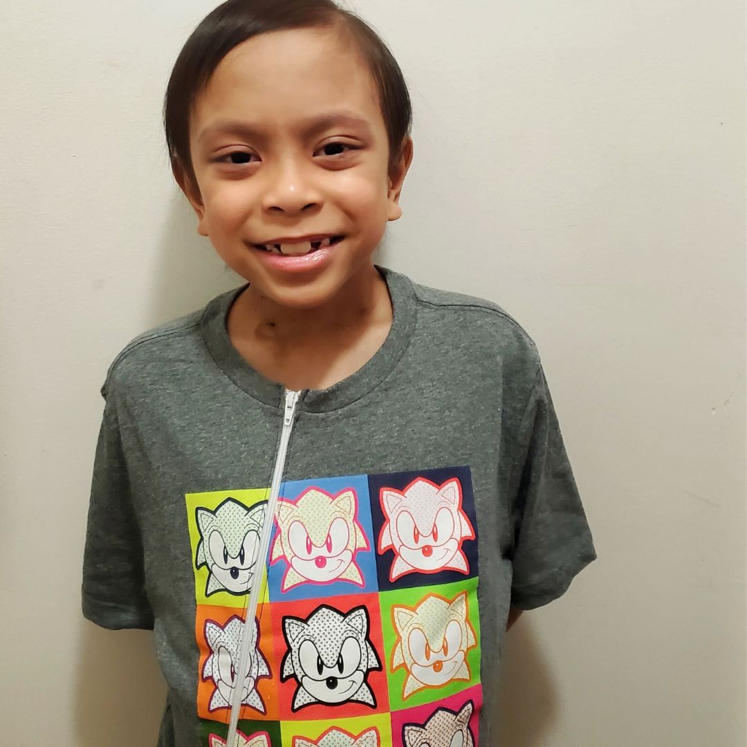 A ten-year-old boy smiling at the camera. He is wearing a Port Shirt with pictures of Sonic the Headgehog on it; the white zipper goes from the collar down the right hand side of the shirt.