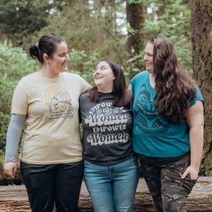 Three young adults at the 2023 She Can retreat (The Club was formerly called She Can). They are stood in a line with their arms round each other's shoulders, looking and smiling at each other. There is a forest of trees in the background.